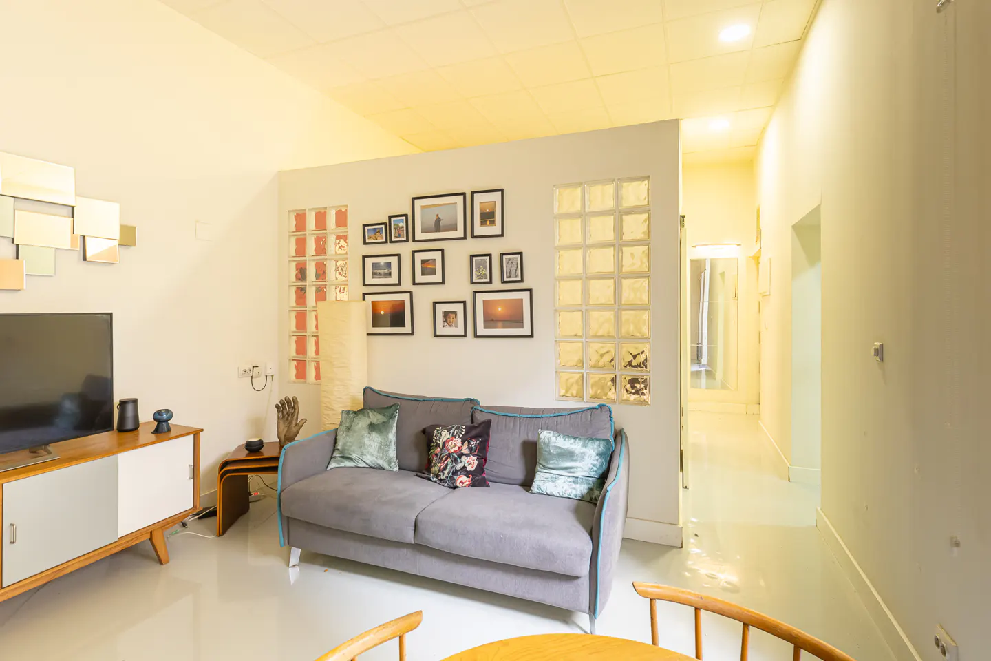 Living room with a gray sofa, pillows, TV, and a gallery wall with photos and glass blocks. Hallway in the background.