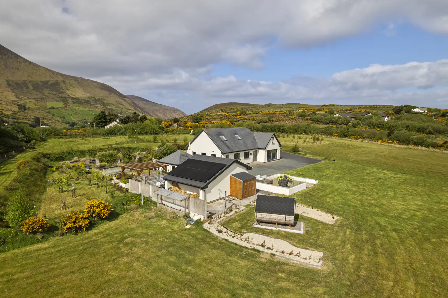 Aerial view of a white house with solar panels, a garden, and a small outbuilding in a scenic, mountainous rural landscape.