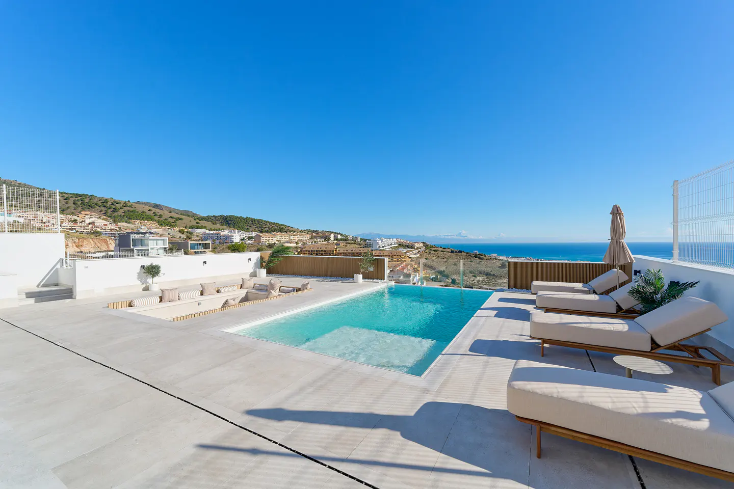 Rooftop pool with lounge chairs and ocean view. Blue sky above, with hills and buildings in the background.