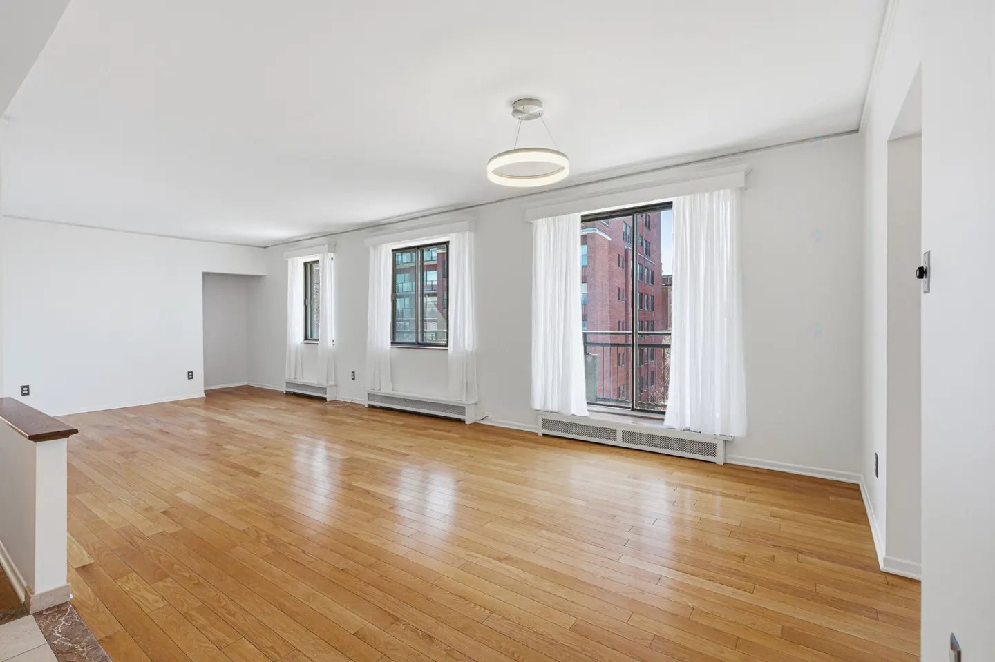 Bright, empty room with hardwood floors, white walls, and three windows with sheer white curtains. A modern light fixture hangs from the ceiling.
