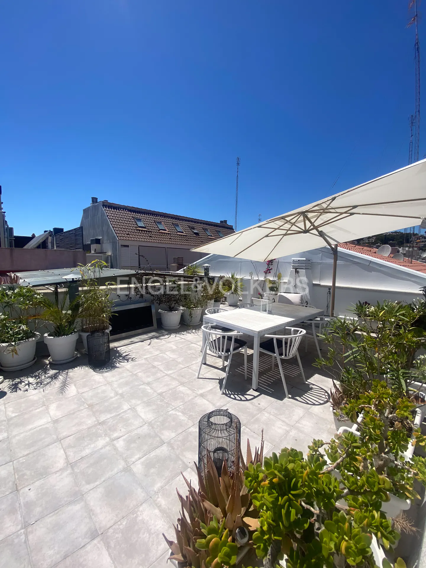 Rooftop terrace with white table, chairs, and umbrella. Potted plants line the perimeter. Blue sky above.