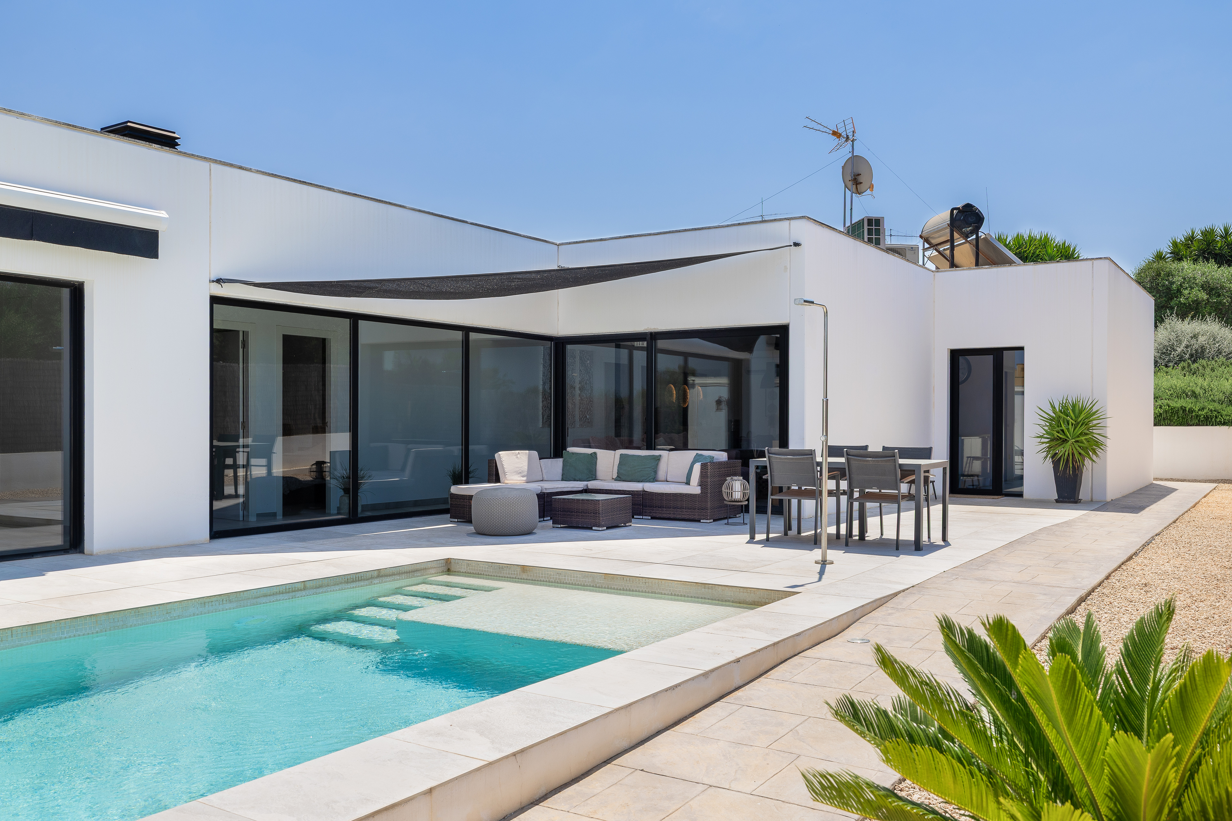 Modern white villa exterior with a swimming pool, patio furniture, and a black shade sail under a clear blue sky.