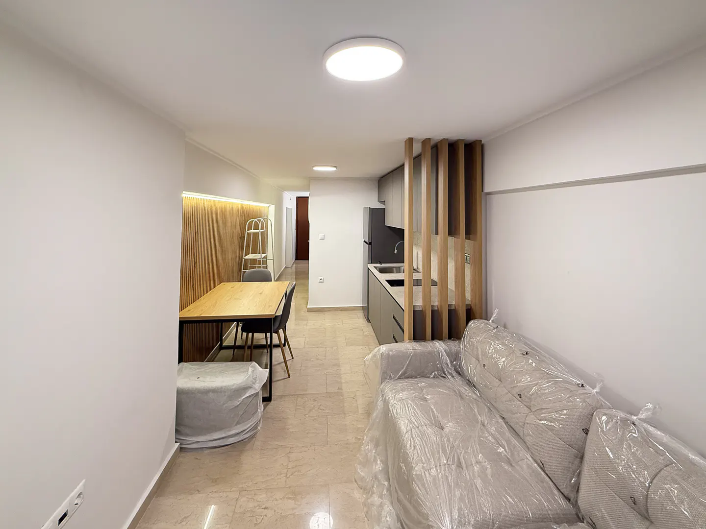 A modern apartment interior with a dining table, kitchen, and sofa covered in plastic. The walls are white, and the floor is beige tile.