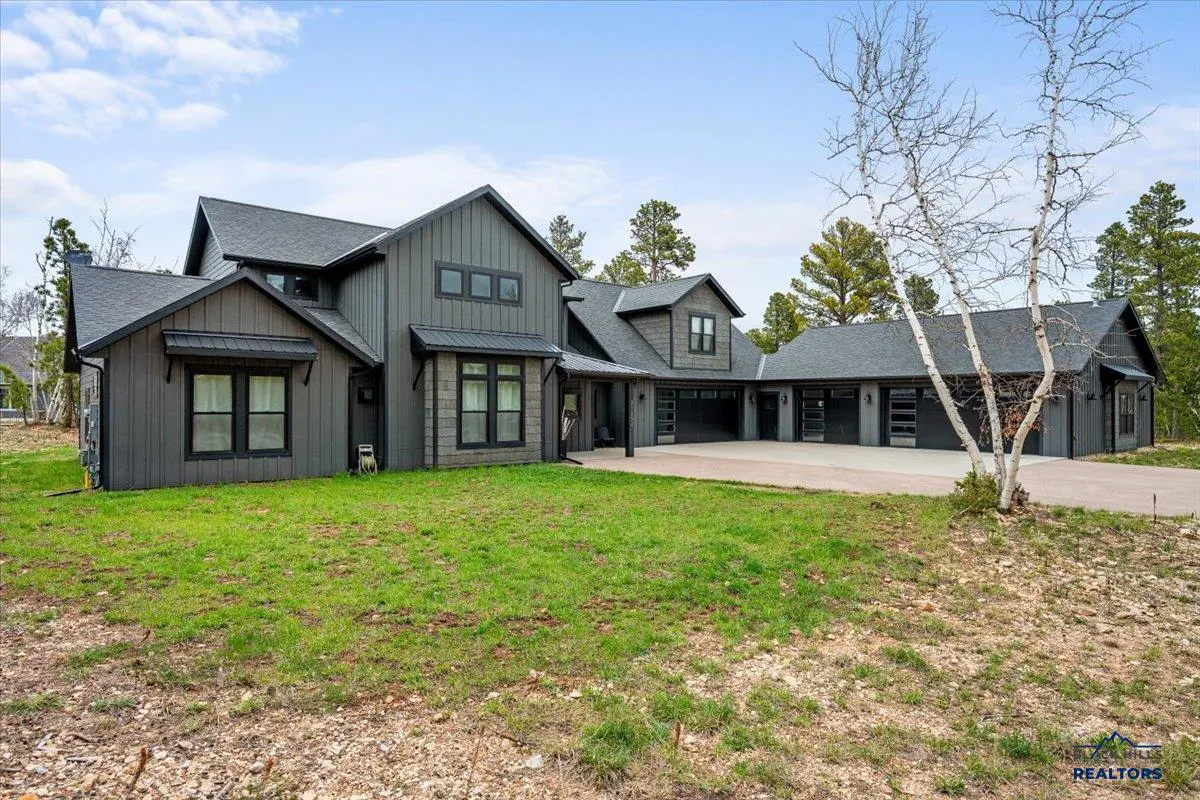 Modern dark gray farmhouse with a large driveway, multi-car garage, and green lawn under a blue sky with scattered clouds.