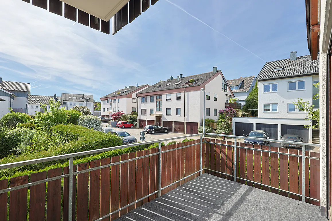 View from a balcony with a brown wood and metal railing, overlooking a street with white houses and parked cars.