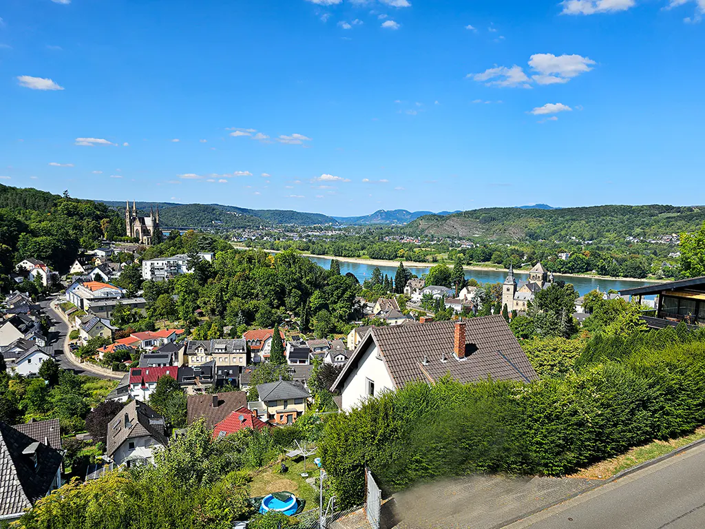 Scenic view of a European town with a river, green hills, and a church under a blue sky with scattered clouds.