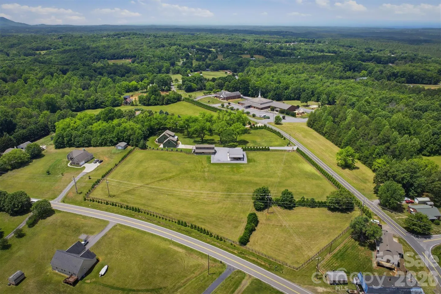 Aerial view of a large, green, grassy lot with a few houses, a church, and a road running through it. The background is a dense forest.