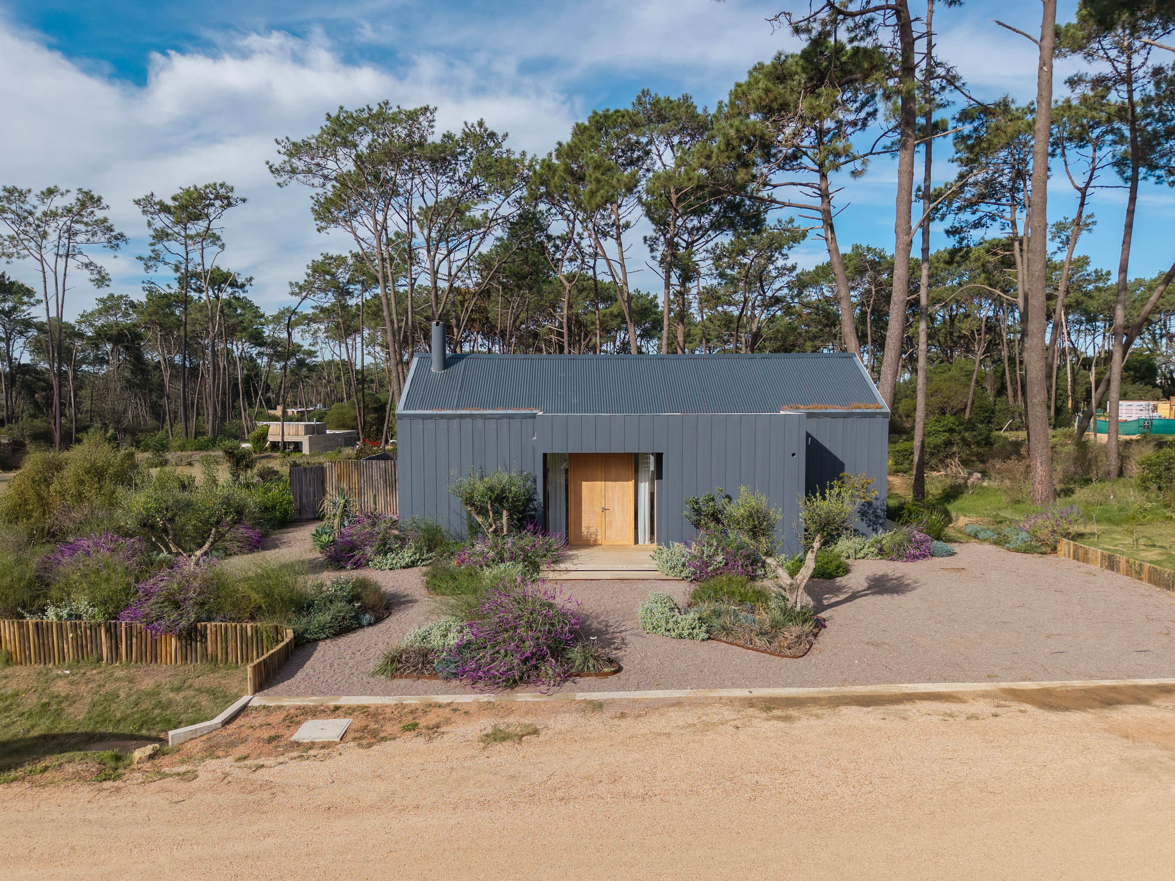 Modern gray house with a metal roof, surrounded by trees and gravel landscaping with purple flowers.