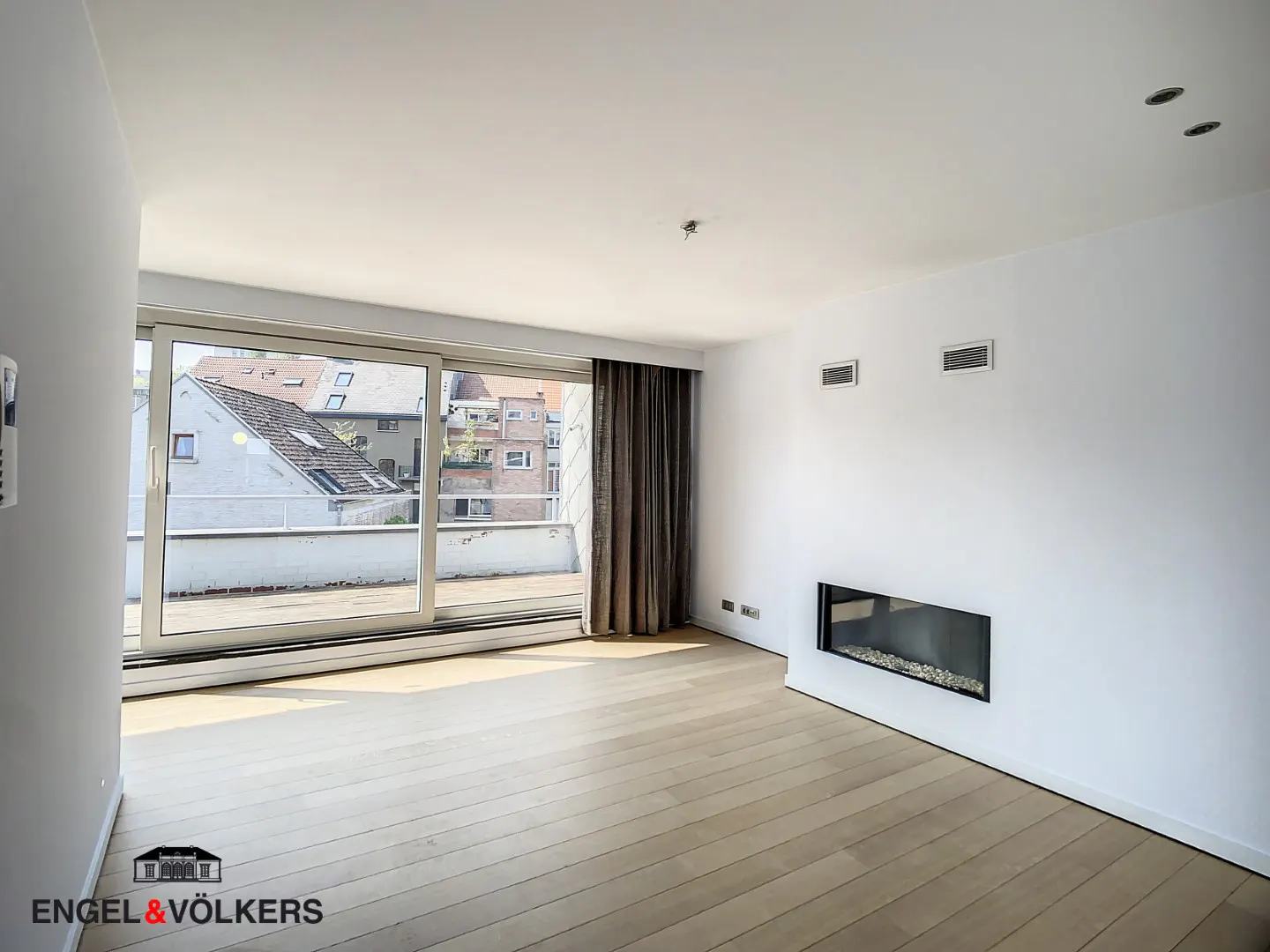 Bright, empty room with light wood floors, white walls, and a modern fireplace. Sliding glass doors lead to a balcony with city views.