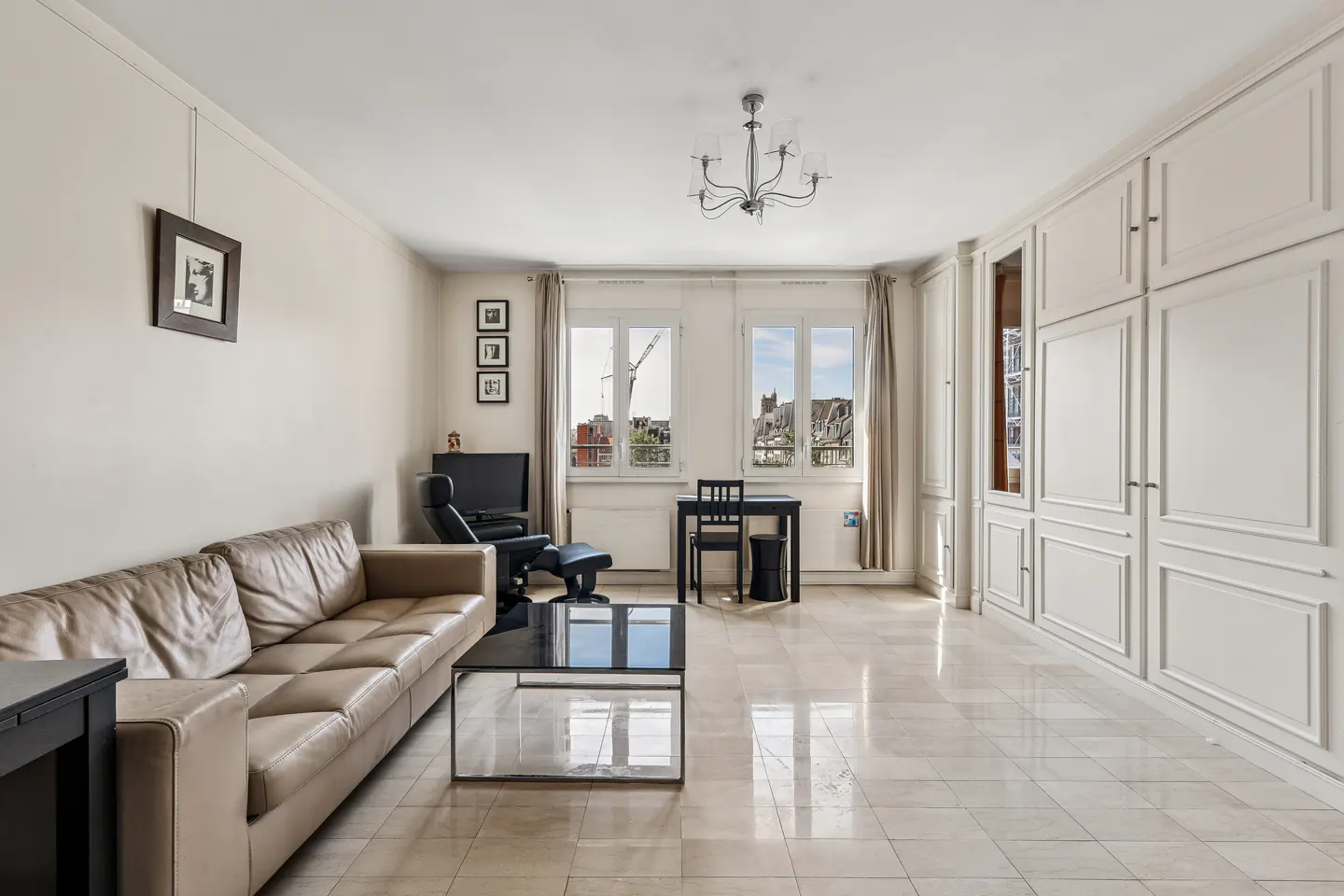 Bright living room with a beige leather sofa, black coffee table, and white built-in cabinets. Windows overlook a city view.
