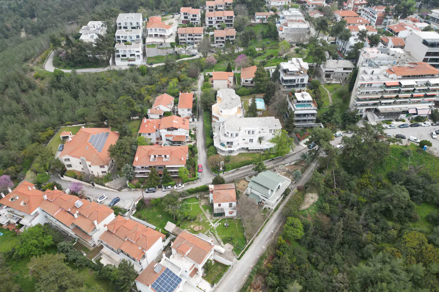 Aerial view of a residential area with houses, trees, and winding roads. The houses have red tile roofs and white walls.