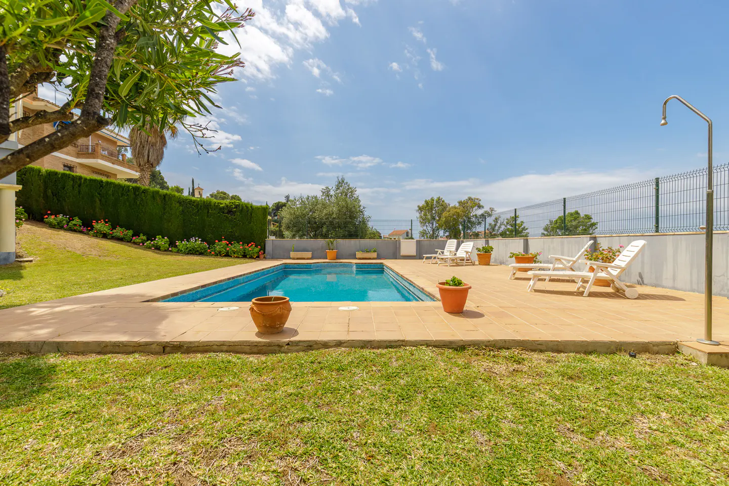 Outdoor pool with blue water, surrounded by a tiled patio, green lawn, and lounge chairs under a sunny sky.
