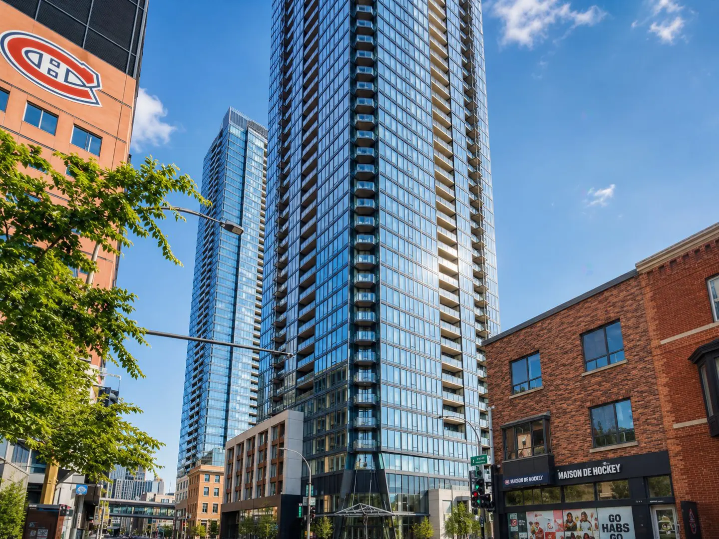 Exterior view of a modern glass skyscraper with balconies, next to a brick building with "Maison de Hockey" sign.