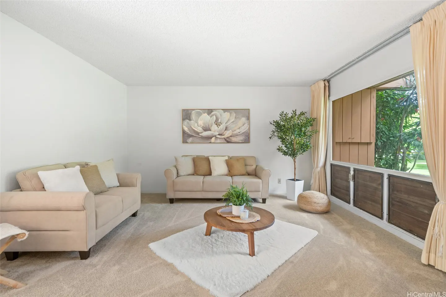 Living room with beige sofas, round wood table on a white rug, and a large window with tan curtains. A floral painting hangs above the sofa.