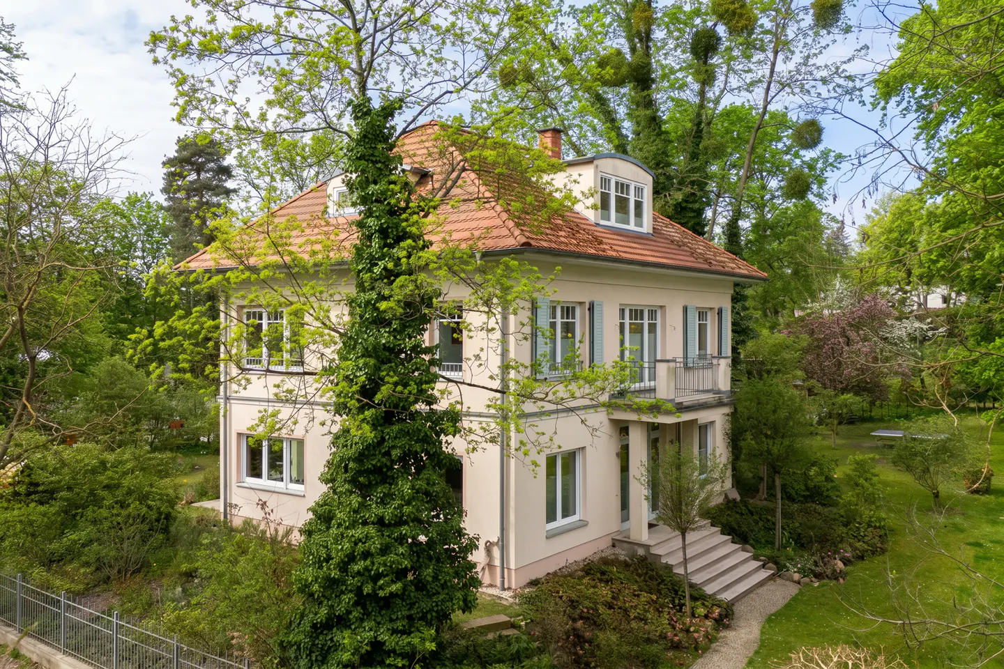 A two-story beige house with a red tile roof, light blue shutters, and a small balcony, surrounded by a lush green garden.