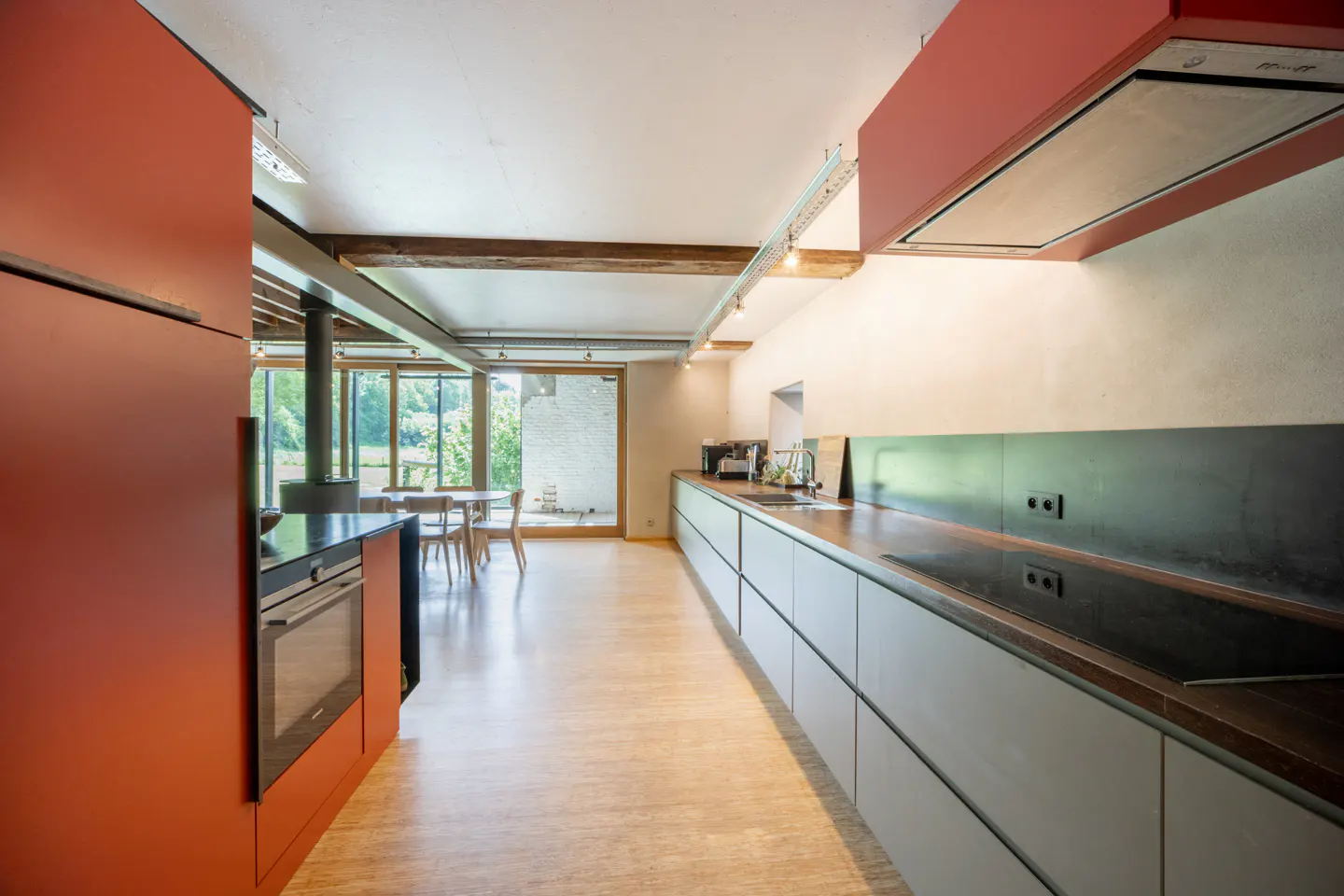 Modern kitchen with red cabinets, grey lower units, wooden floors, and a dining area overlooking a garden.