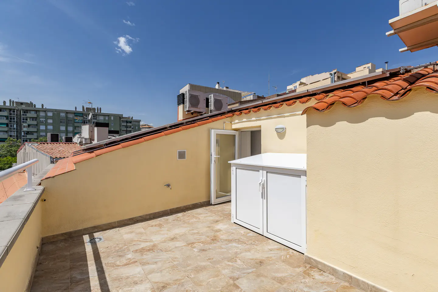 A rooftop terrace with beige tile flooring, a white storage cabinet, and a view of city buildings under a blue sky.
