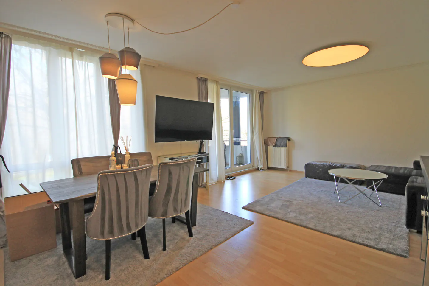 A modern living room with a dining table, gray chairs, a TV, and a gray rug on a wood floor.