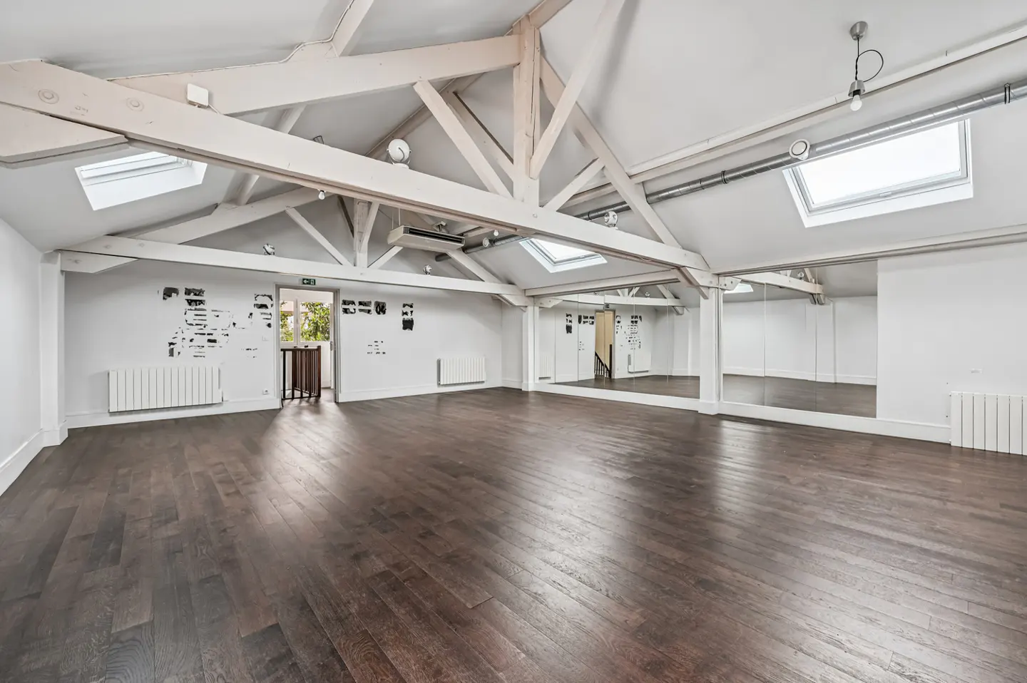 Bright, empty studio with dark wood floors, white walls, and exposed white beams. A wall of mirrors reflects the room.