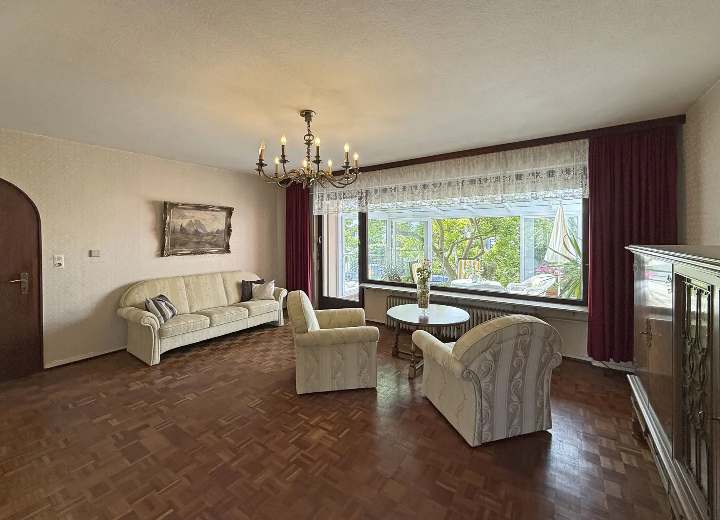 A living room with parquet floors, a cream sofa, two armchairs, and a round table by a large window. A chandelier hangs from the ceiling.