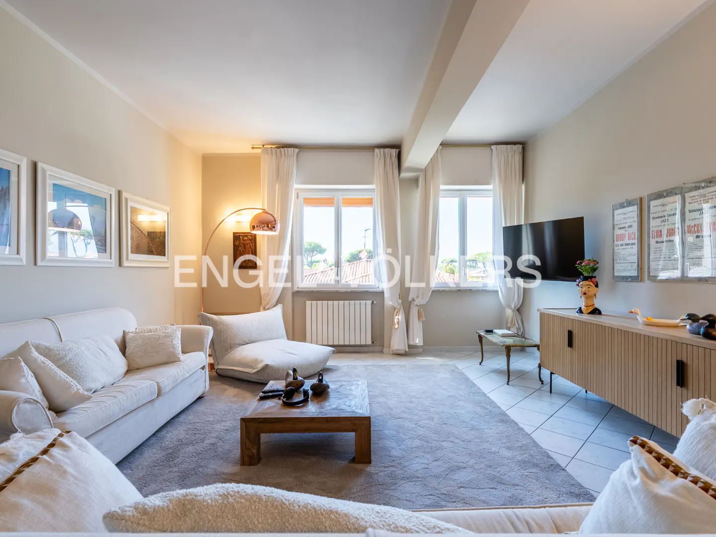 Living room with white sofas, wood coffee table, and gray rug. Windows with white curtains let in natural light. TV and cabinet on the right.