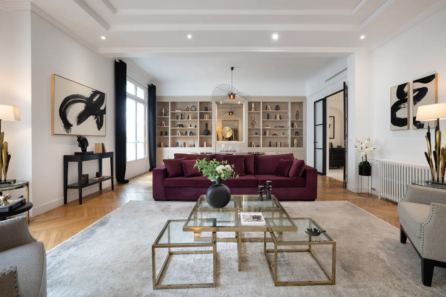 Elegant living room with a deep burgundy sofa, glass coffee tables, built-in bookshelves, and modern abstract art.