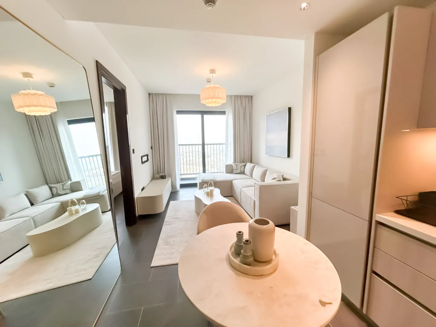 Modern apartment interior with a round dining table in the foreground, a large mirror, and a living area with a sofa.