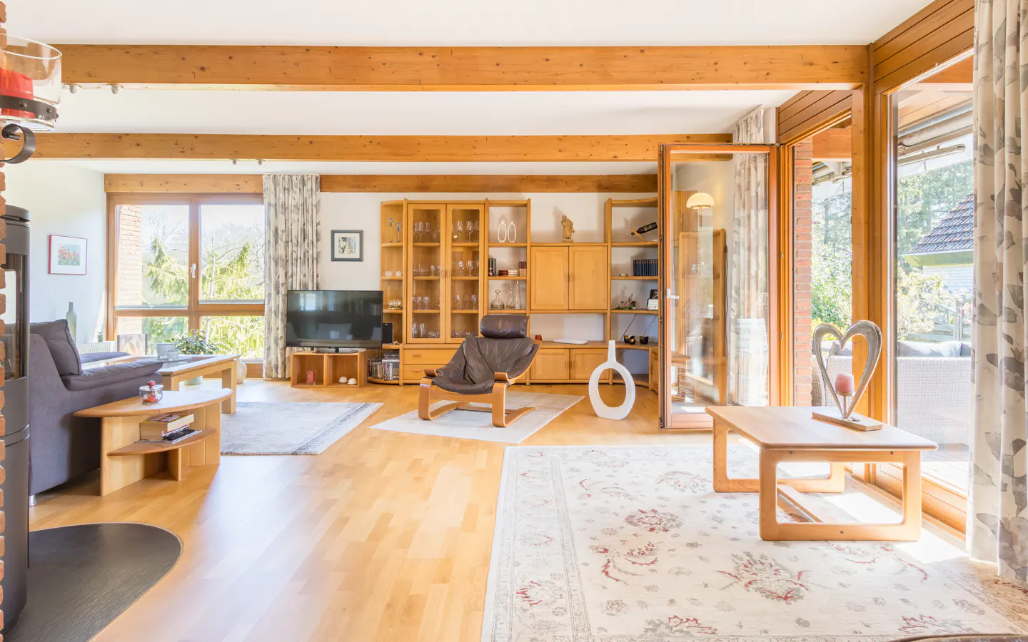 Bright living room with hardwood floors, exposed beams, and large windows. A brown leather chair sits near a wooden shelving unit.