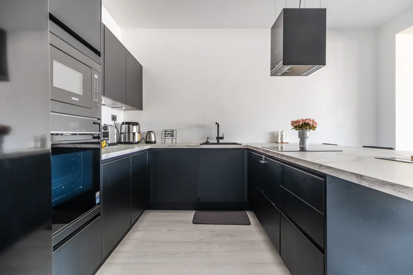 A modern kitchen with dark blue cabinets, white marble countertops, and stainless steel appliances. A black range hood hangs above the island.