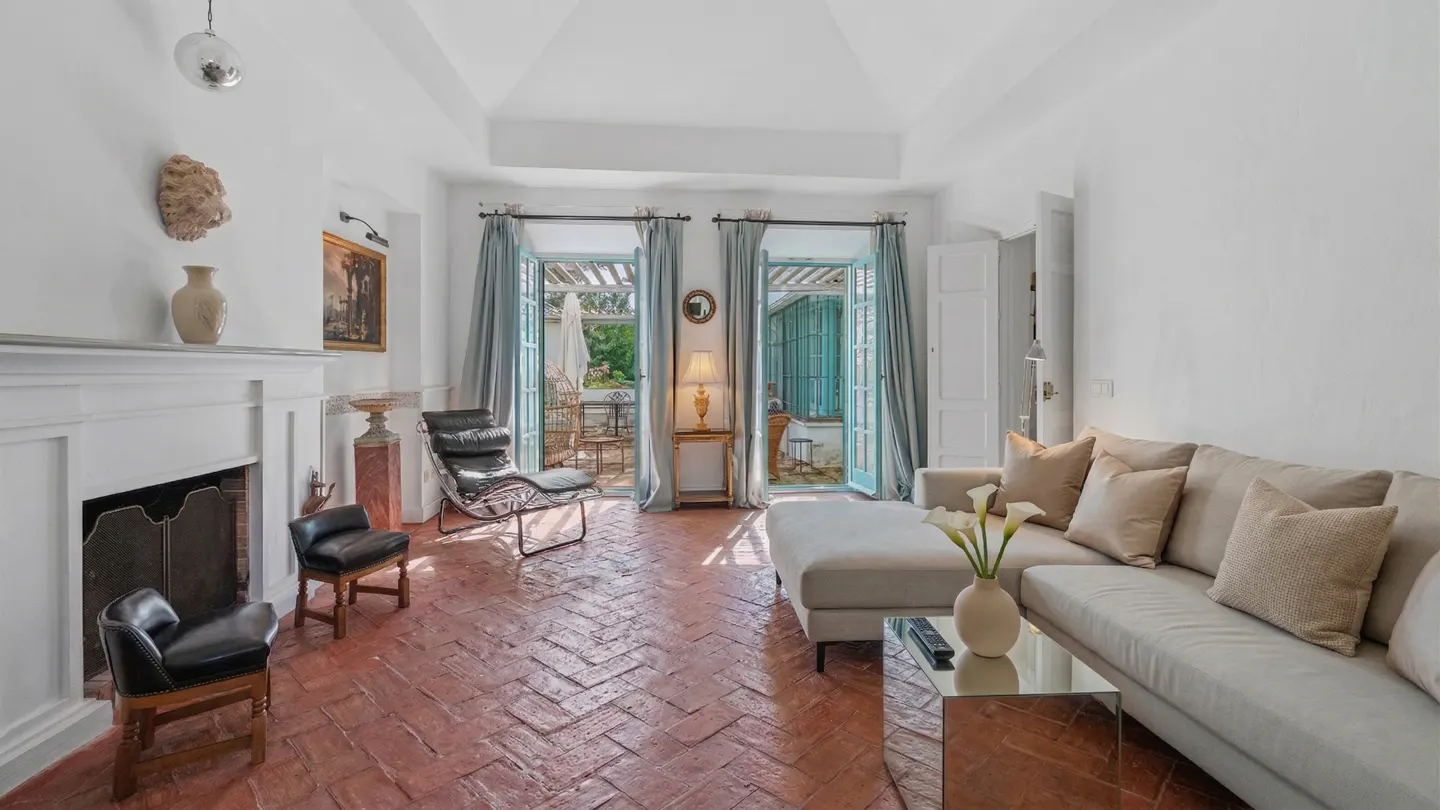 Bright living room with white walls, terracotta floors, and a beige sectional sofa. French doors with blue curtains lead to a patio.