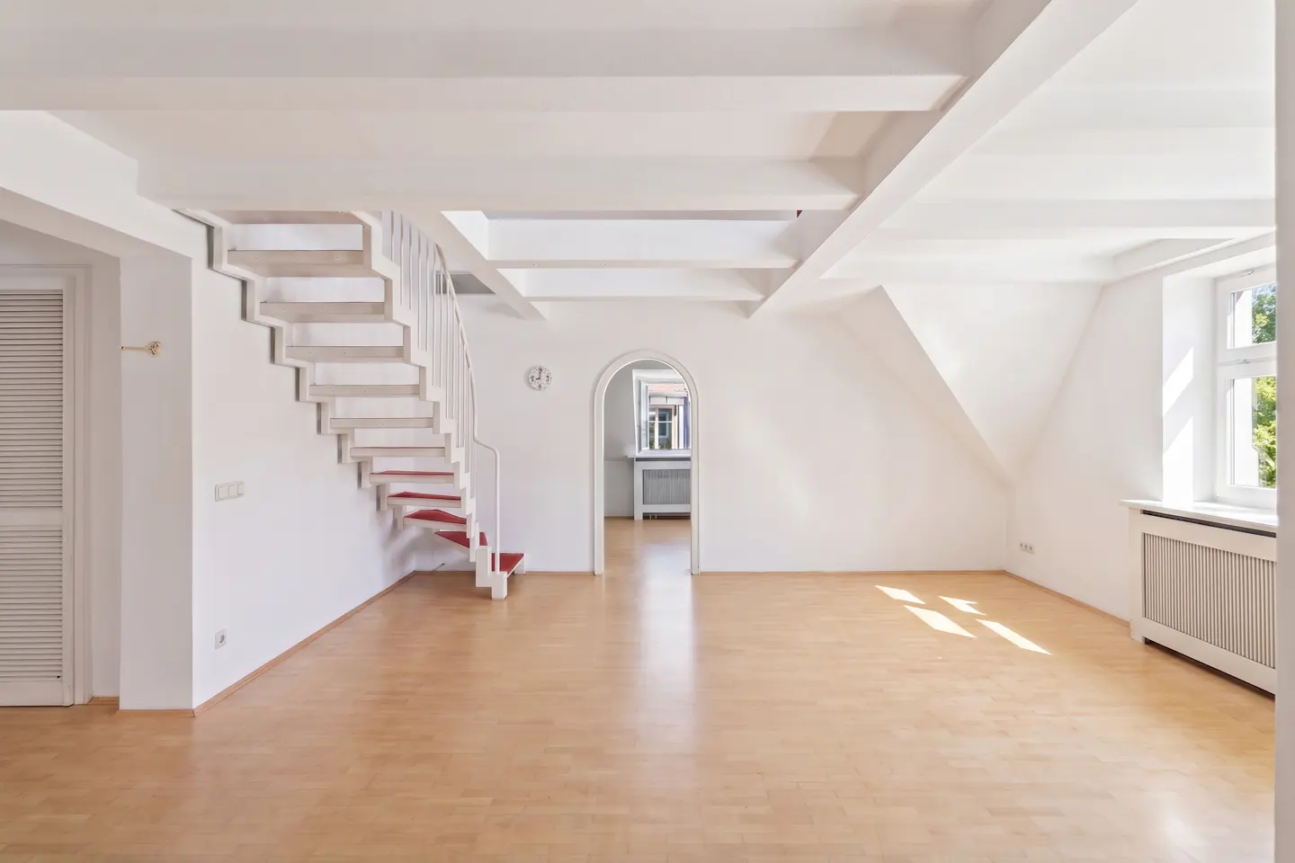 Bright, empty room with hardwood floors, white walls, and exposed beams. A white staircase with red treads leads up. Arched doorway in the back.