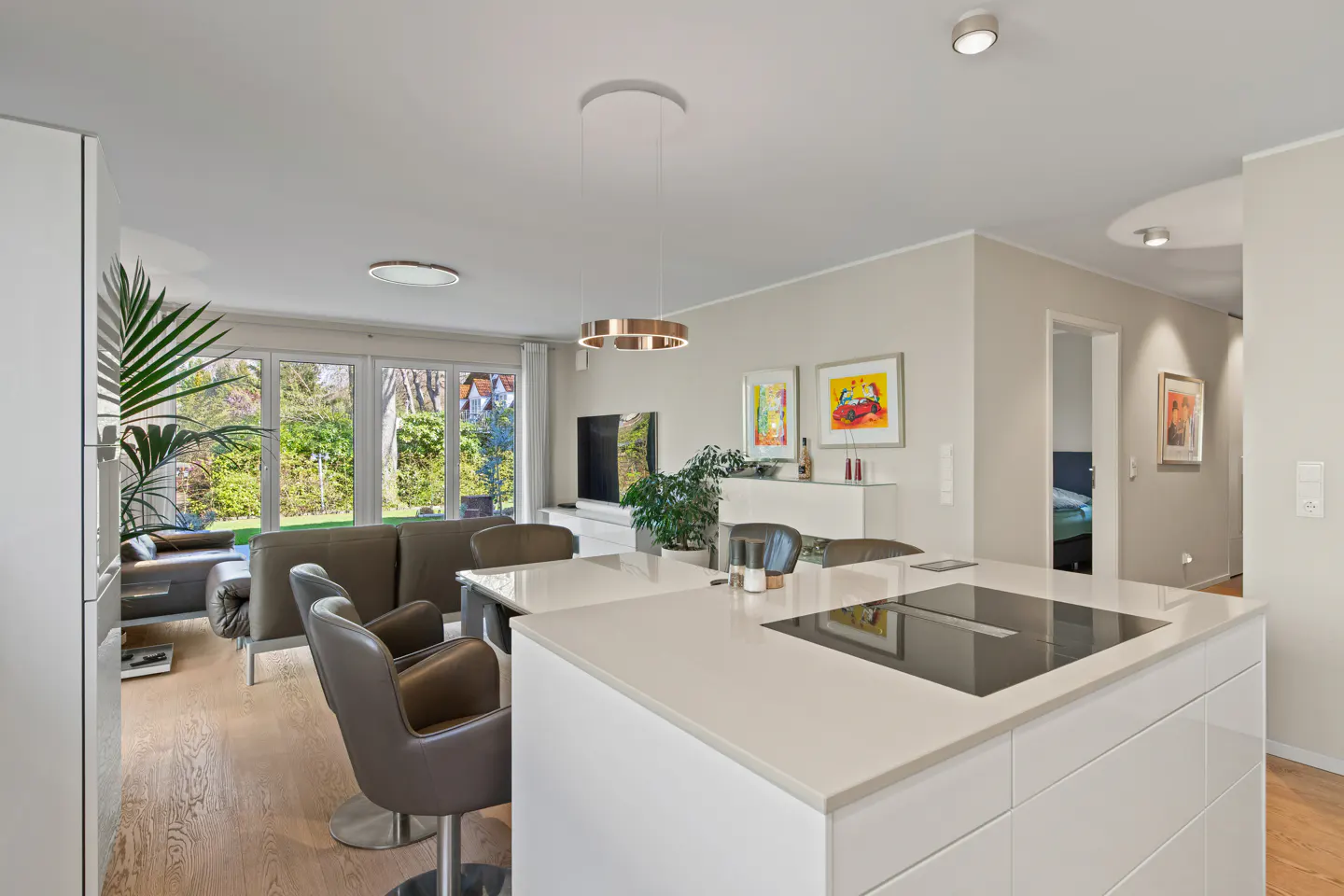 Bright, modern open-plan living space with white kitchen island, grey chairs, and a view to a green garden through large windows.