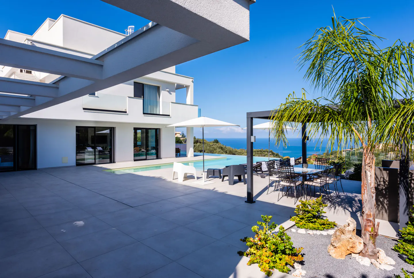 Modern white house with pool, patio, and ocean view. Palm tree and outdoor furniture add to the luxurious setting.