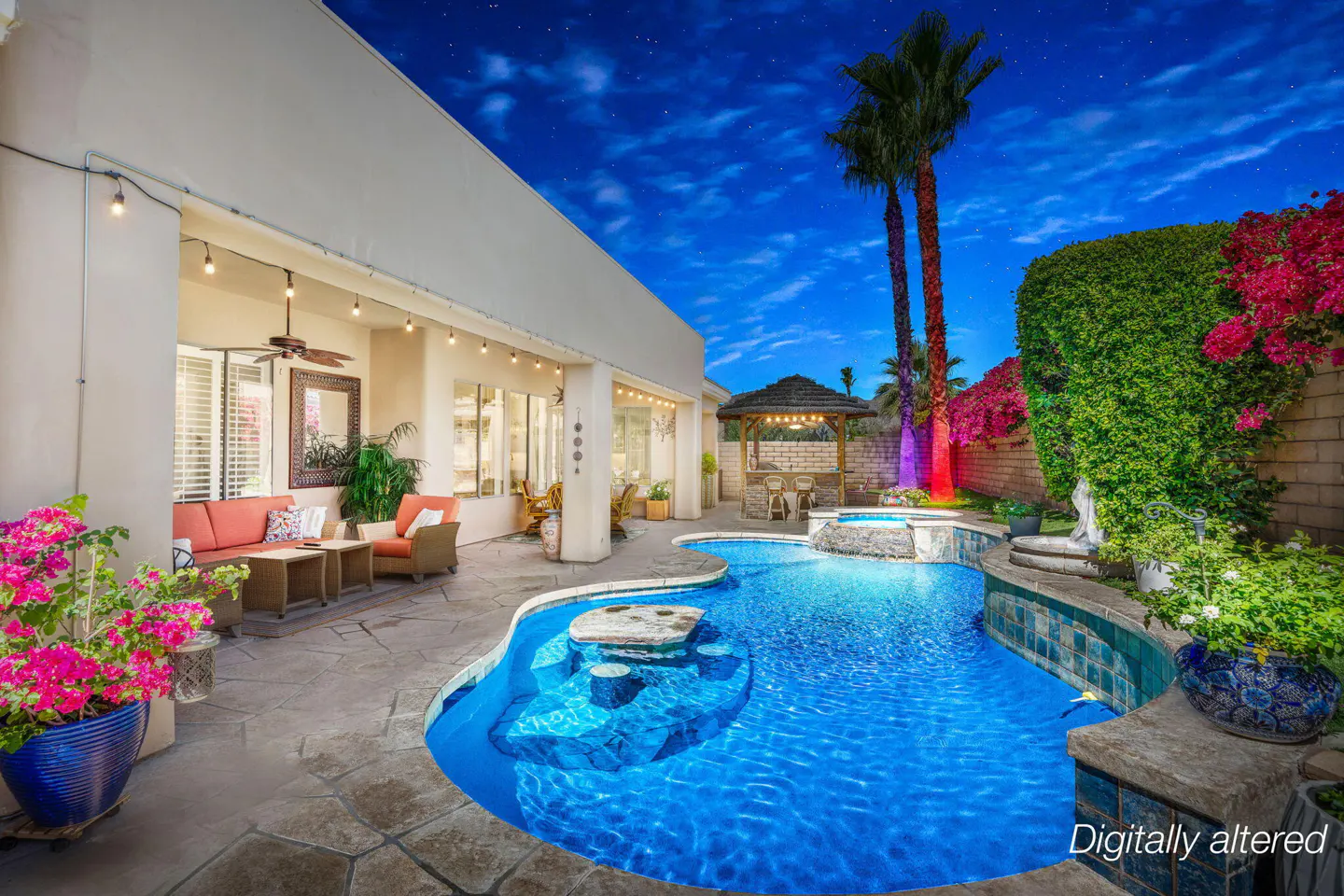 Backyard at night with a blue pool, stone patio, covered seating area, palm trees, and vibrant pink bougainvillea.