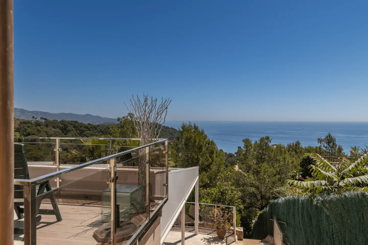 Balcony view with glass railings overlooking the ocean and green trees under a clear blue sky.