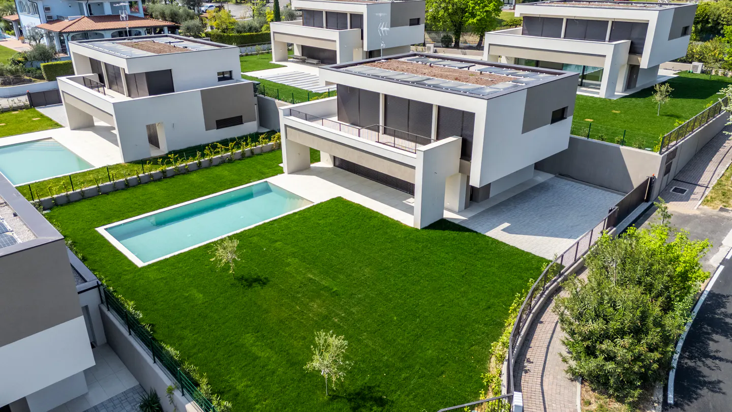 Aerial view of modern white houses with pools, surrounded by green lawns and trees.