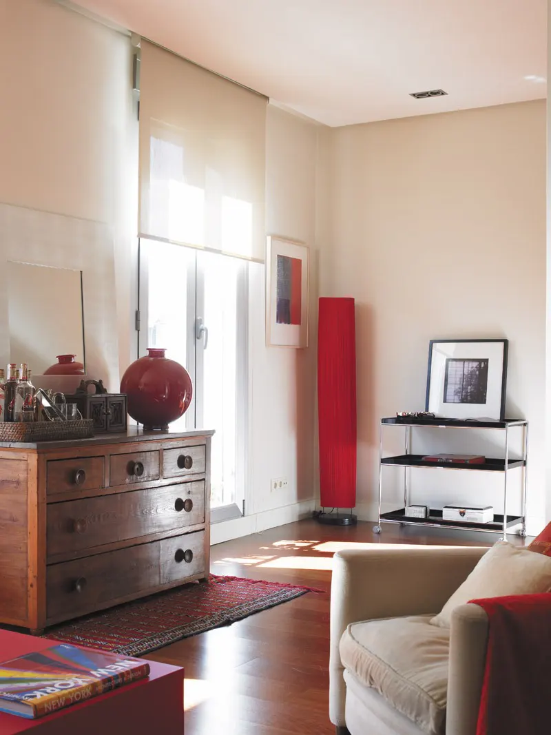Living room with a wooden dresser, red accents, a beige sofa, and a black and silver TV stand. Natural light filters through the window.