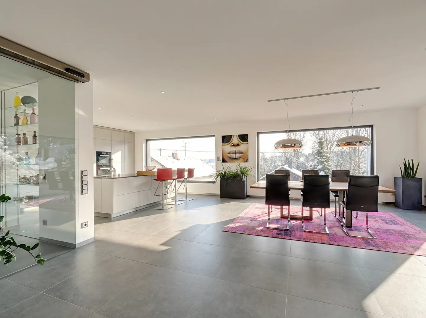 Modern open-plan dining and kitchen area with gray tile floors, a pink rug, black chairs, and large windows with snowy views.