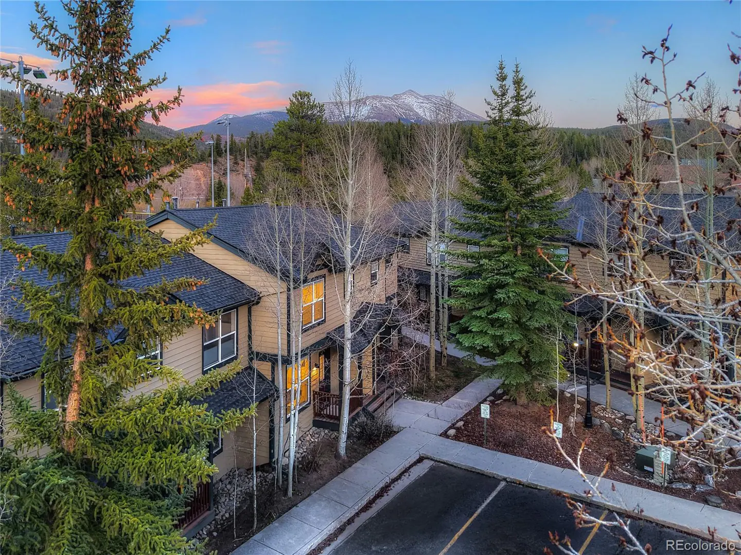 Aerial view of tan townhouses with dark roofs, surrounded by trees. A snow-capped mountain is visible in the background under a blue and pink sky.