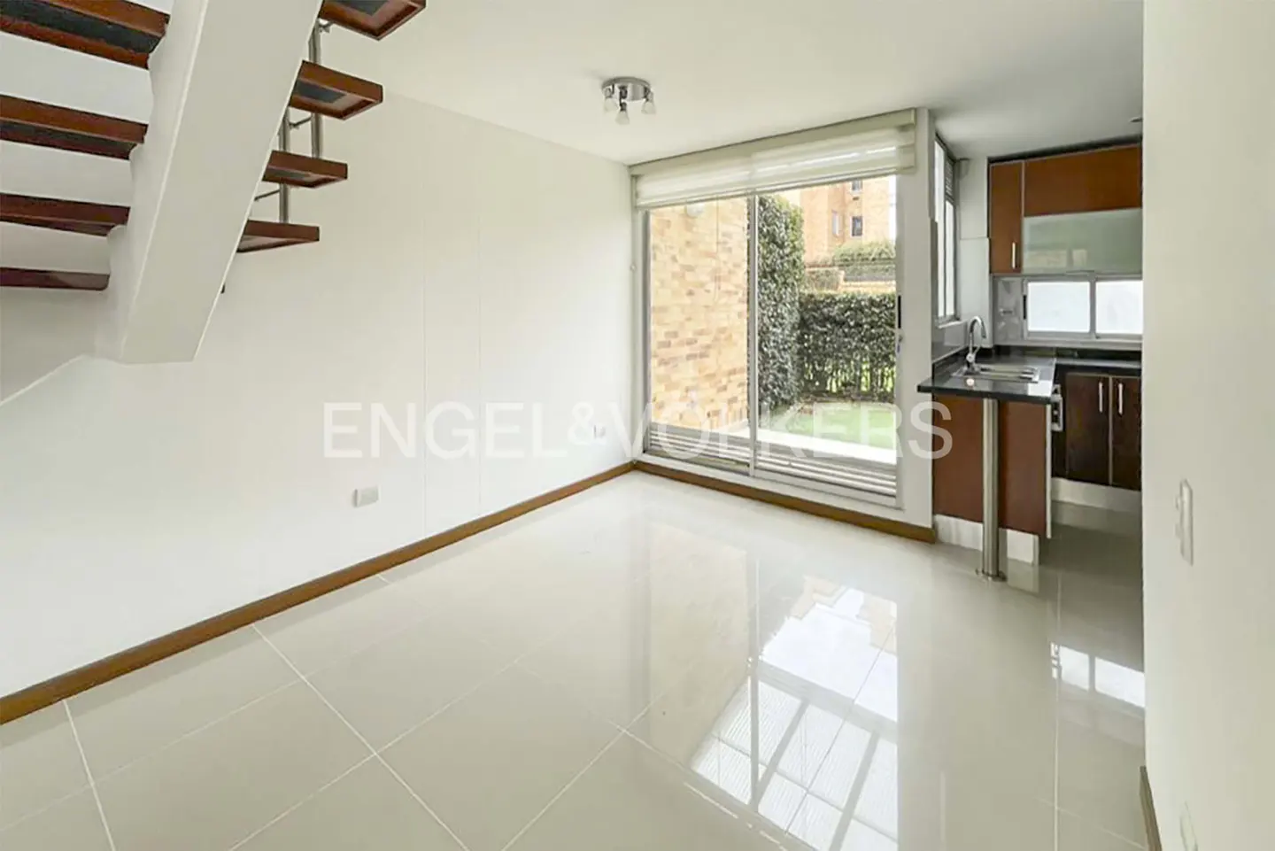 Bright, modern apartment interior with white walls, tile floor, and wooden stairs. A sliding glass door leads to a green outdoor space. A compact kitchen area is visible.