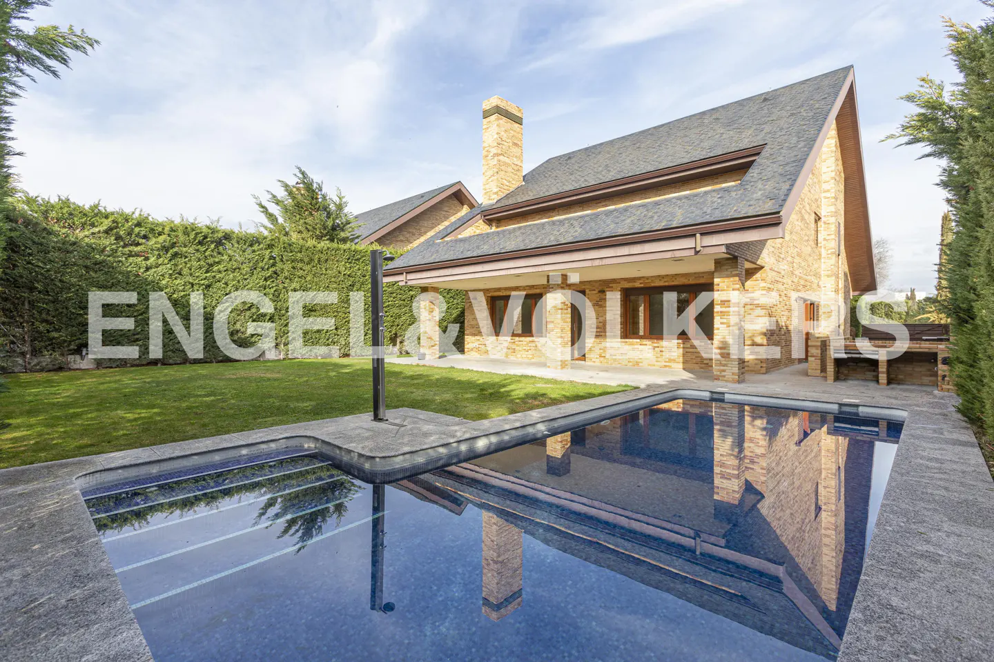 Exterior view of a tan brick house with a pool, green lawn, and tall hedges under a partly cloudy sky.