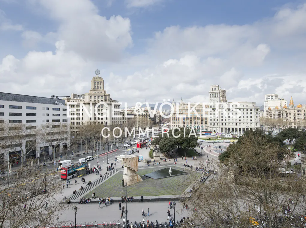 Aerial view of Plaça de Catalunya in Barcelona, Spain, with buildings, trees, a fountain, and people. Blue sky with clouds.