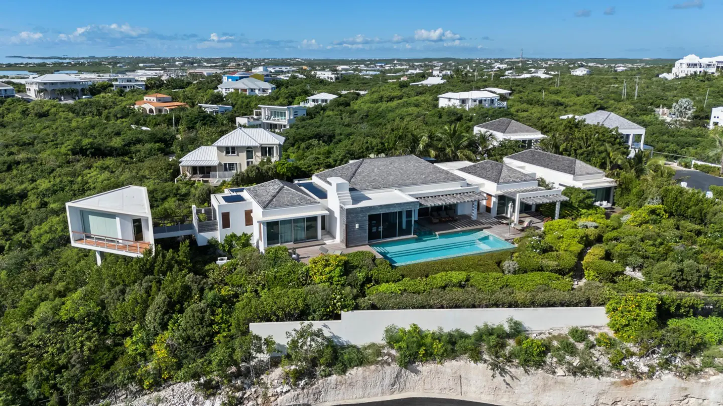 Aerial view of a modern white house with a pool, surrounded by lush green trees and other houses in the background.