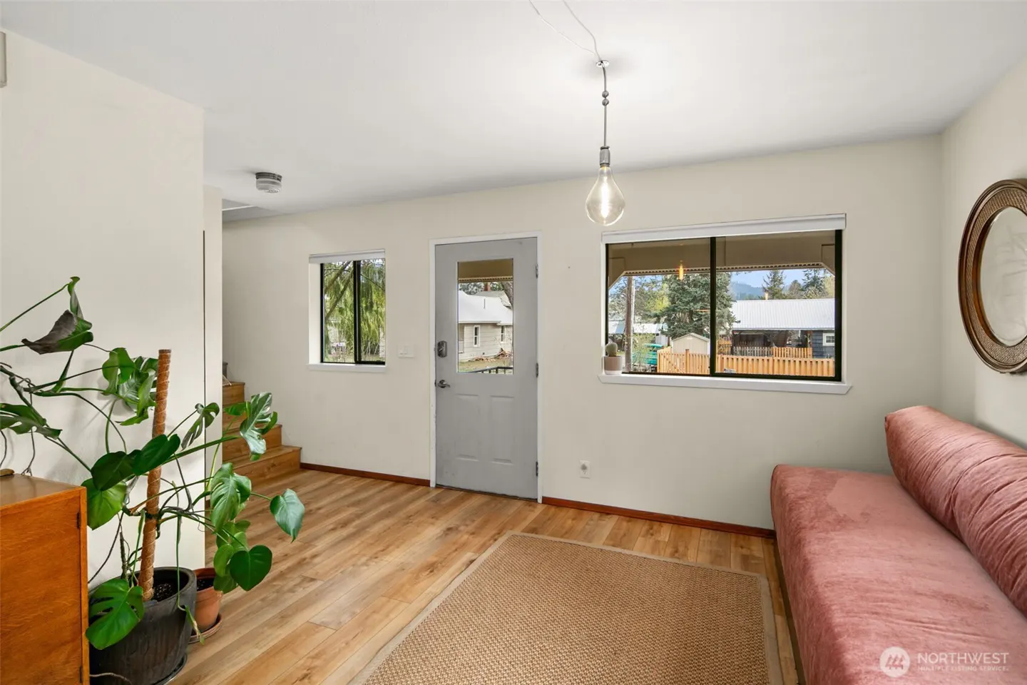 Living room with wood floors, a pink sofa, and a jute rug. A gray door and windows look out to a yard. A monstera plant sits near stairs.