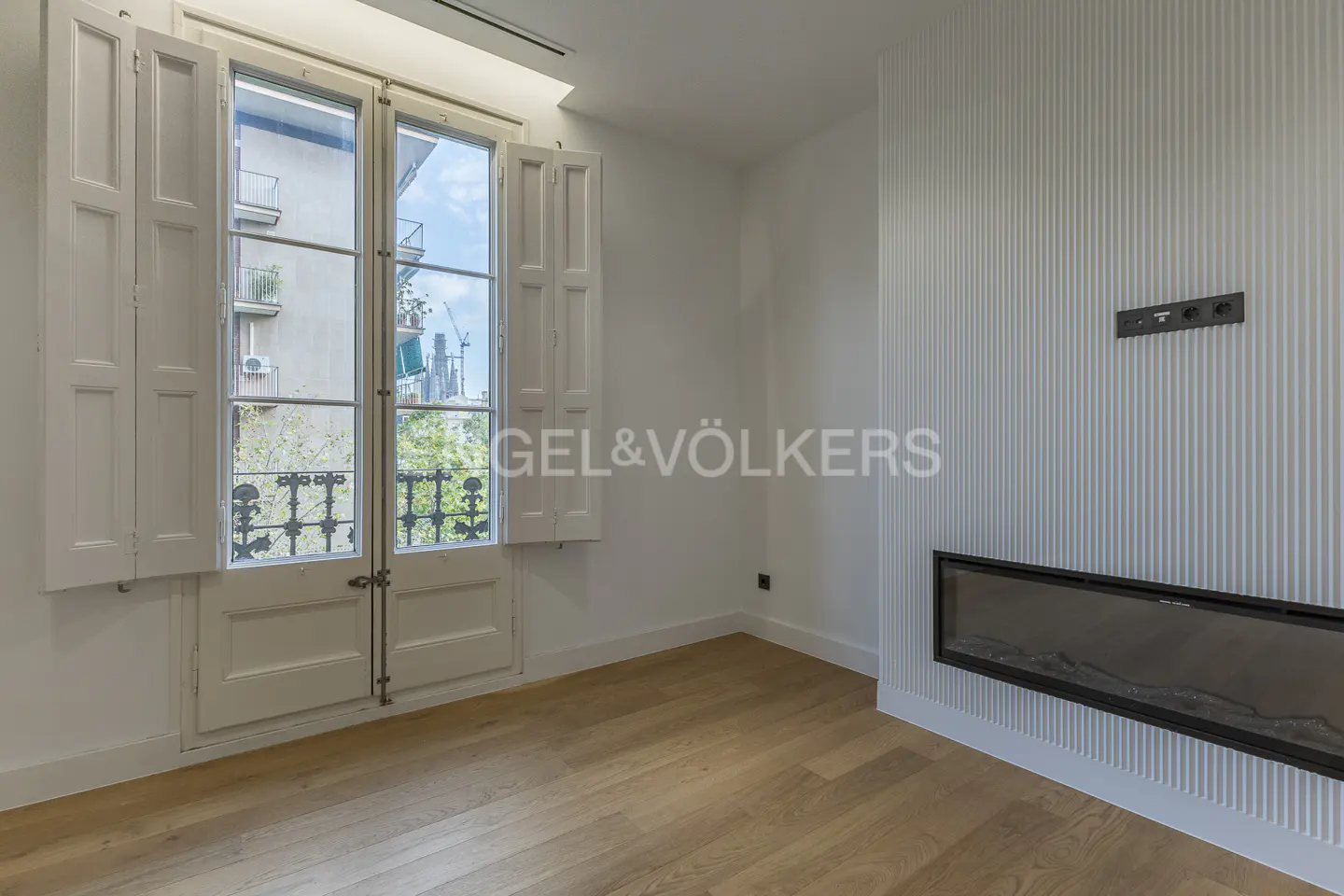 Bright room with wood floors, white walls, and a modern fireplace. Open white shutters reveal a city view.