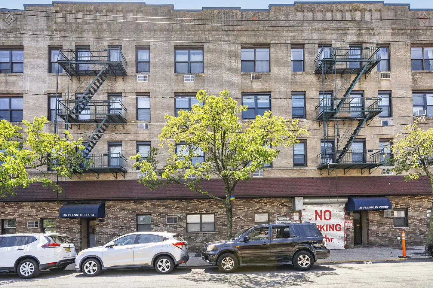 A multi-story tan brick apartment building with black fire escapes, green trees, and parked cars in front.