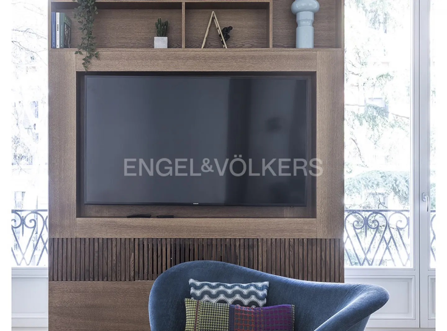 Living room with a large TV mounted on a wooden wall unit, a blue chair with pillows, and a window with a view.