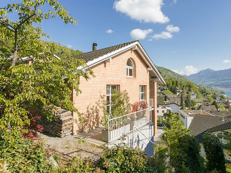 Exterior view of a salmon brick house with a white balcony, green trees, and a mountain view under a blue sky.