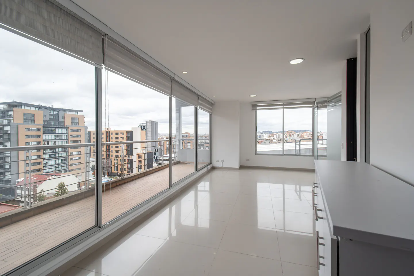 Bright, empty apartment with white walls, tile floors, and large windows overlooking a city skyline and balcony.