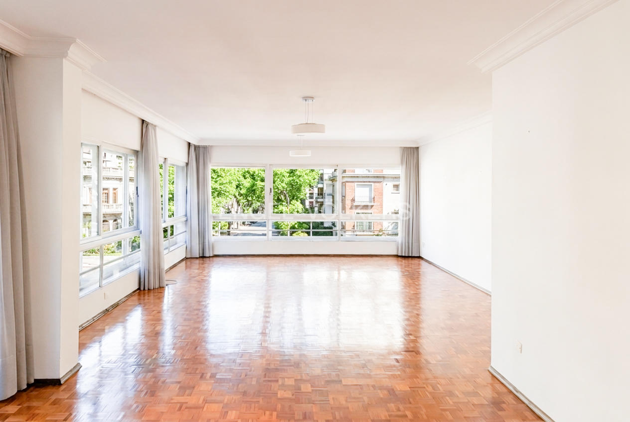 Bright, empty living room with polished parquet wood floors, white walls, and large windows overlooking green trees. [AI-generated text]